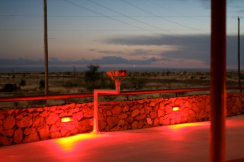 A desert landscape at dusk, featuring a stone wall and a glowing red light, with distant clouds on the horizon.