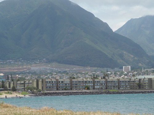 A coastal view featuring a low-rise building along the shore, with mountains rising in the background under a cloudy sky.