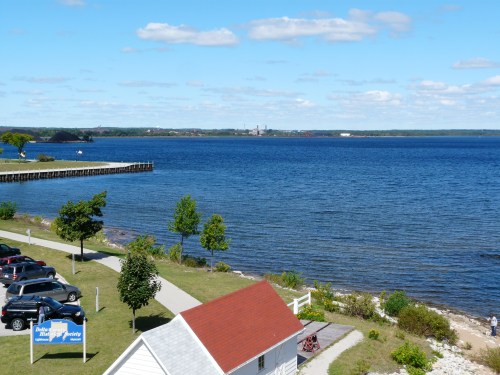 A serene lakeside view with a blue sky, distant industrial buildings, and a small white house near the water.