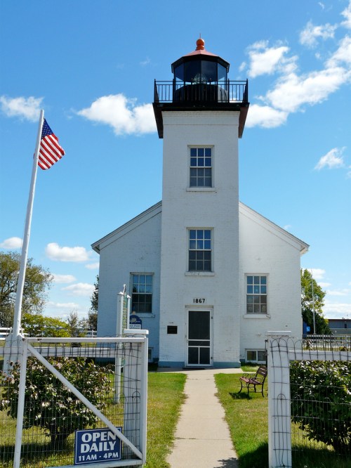 A white lighthouse with a red roof, surrounded by grass, a flagpole, and a sign indicating open hours.