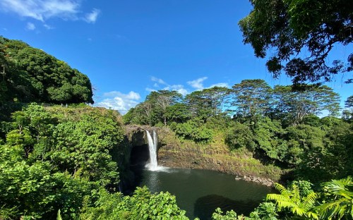 A lush green landscape featuring a waterfall cascading into a serene pool, surrounded by trees and blue sky.