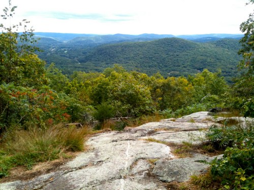 A scenic view from a rocky outcrop, overlooking lush green hills and valleys under a cloudy sky.