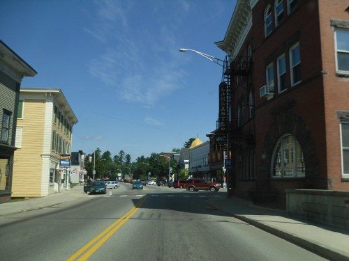 A sunny street scene with buildings on either side, cars parked, and a clear blue sky overhead.