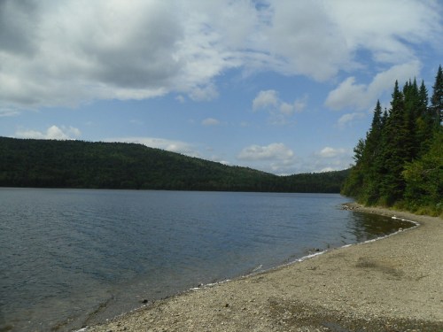 A serene lake surrounded by lush green hills and a sandy shore under a partly cloudy sky.