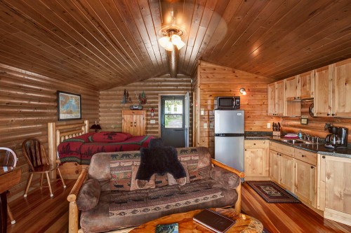 Cozy wooden cabin interior with a bed, sofa, kitchen area, and rustic decor. Warm lighting enhances the inviting atmosphere.