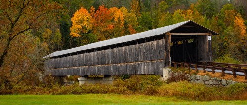 A rustic wooden covered bridge surrounded by vibrant autumn foliage and greenery.