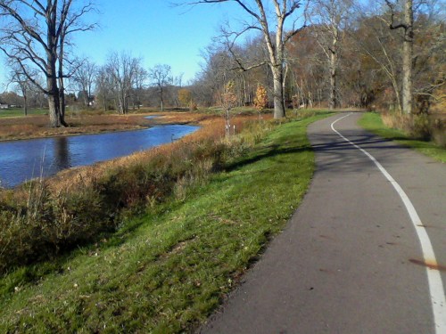 A scenic pathway alongside a calm river, surrounded by trees and autumn foliage under a clear blue sky.