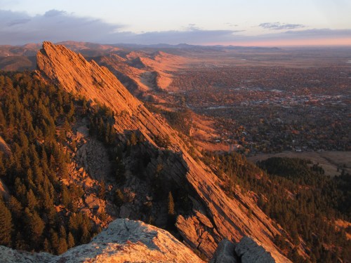 Sunset over a rugged mountain peak, casting warm light on rocky cliffs and a valley below, with distant hills in the background.