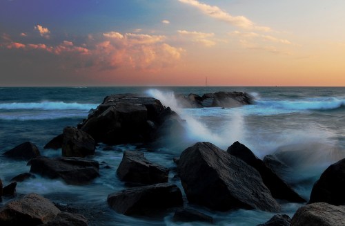 Waves crash against rocky shore at sunset, with a colorful sky and distant sailboat on the horizon.