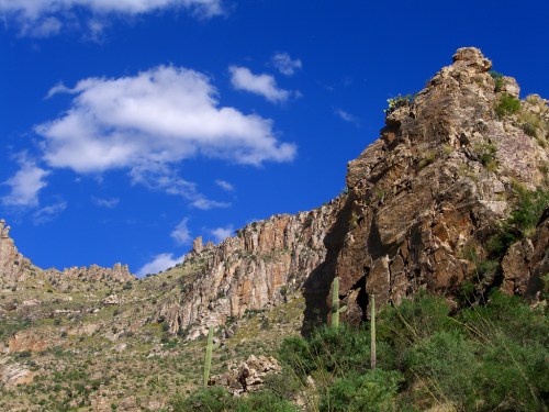 A rugged mountain landscape with rocky cliffs, green vegetation, and a bright blue sky with scattered clouds.
