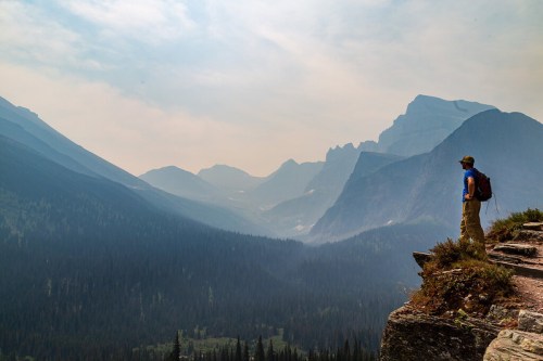 A hiker stands on a rocky ledge, gazing at misty mountains and a lush valley below.
