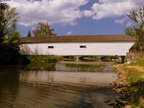A white covered bridge spans a calm river, surrounded by trees and a blue sky with fluffy clouds.