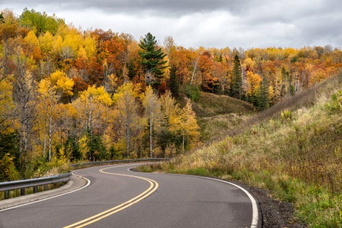 A winding road through a forest with vibrant autumn foliage under a cloudy sky.