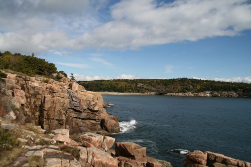 Rocky coastline with calm waters, under a blue sky with scattered clouds and green hills in the background.