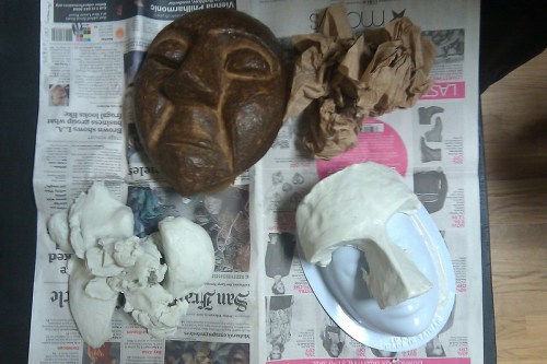 A wooden mask, crumpled paper, and sculpted materials on a newspaper-covered table.