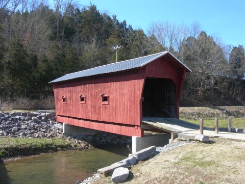 A red covered bridge spans a small stream, surrounded by trees and grassy areas.