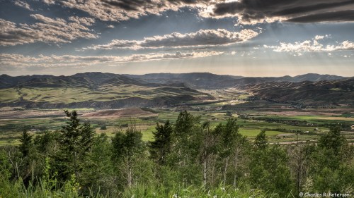 A panoramic view of rolling green hills and valleys under a dramatic sky with rays of sunlight breaking through clouds.