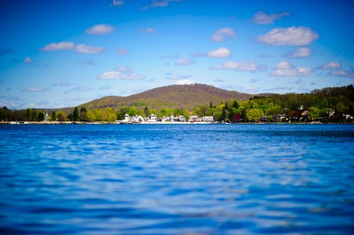 A serene lake scene with a distant shoreline, green hills, and a blue sky dotted with fluffy clouds.