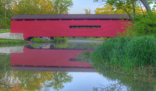 A red covered bridge reflected in calm water, surrounded by lush greenery and trees in the background.