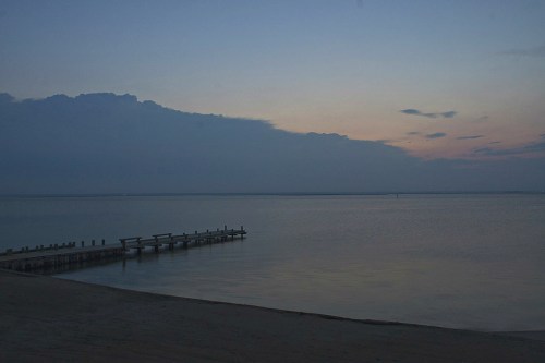 A calm beach at dusk with a wooden pier extending into the water, under a soft, cloudy sky.