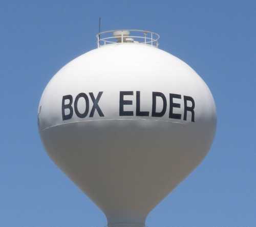 Water tower with the words "BOX ELDER" printed in bold black letters against a clear blue sky.