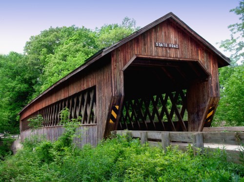 A wooden covered bridge with a sign reading "State Road," surrounded by lush greenery and trees.