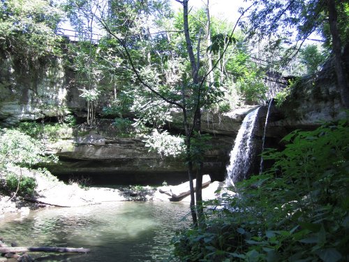 A serene waterfall cascades into a clear pool, surrounded by lush greenery and rocky cliffs.