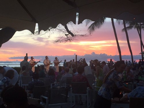 A sunset view with musicians performing on stage, surrounded by an audience at a beachside venue.