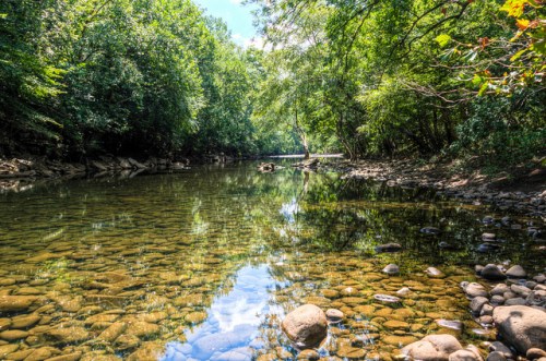A serene river scene with clear water, smooth stones, and lush green trees reflecting in the water under a blue sky.