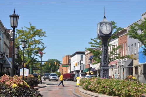 A sunny street scene featuring a clock tower, shops, and a pedestrian walking among colorful flowers.
