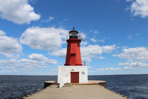 A red lighthouse stands on a pier, surrounded by water under a blue sky with fluffy clouds.