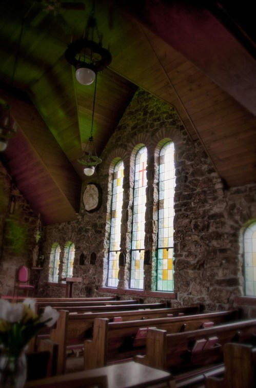Interior of a stone church with colorful stained glass windows and wooden beams, featuring wooden pews and soft lighting.