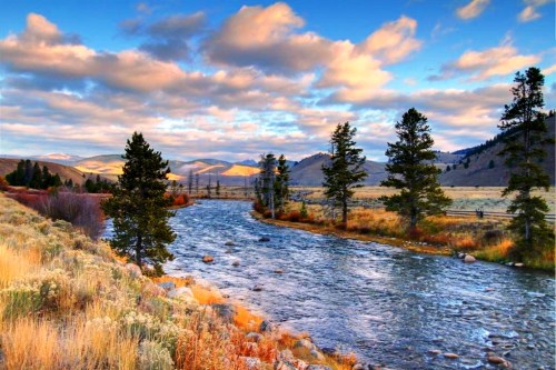 A serene river flows through a landscape of trees and mountains under a colorful sky with fluffy clouds.