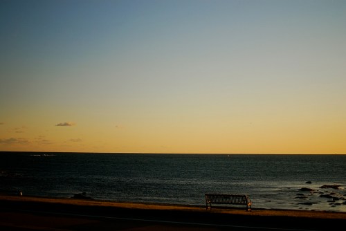 A serene coastal scene at sunset, featuring calm waters and a solitary bench overlooking the horizon.