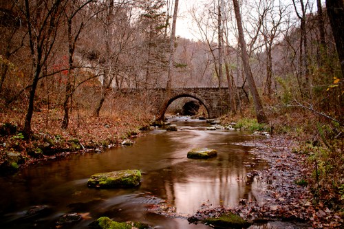 A serene stream flows under a stone bridge, surrounded by trees and autumn foliage.