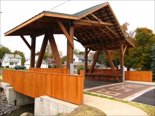 A wooden pavilion with a sloped roof, supported by large beams, set in a residential area with houses in the background.