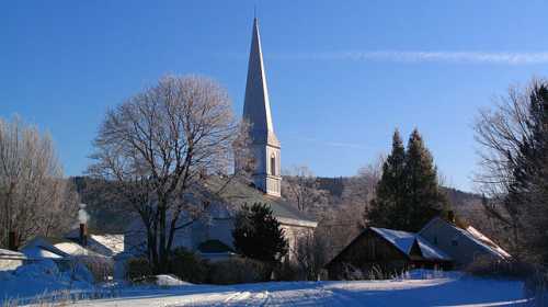 A snowy landscape featuring a church with a tall spire, surrounded by trees and a few buildings under a clear blue sky.