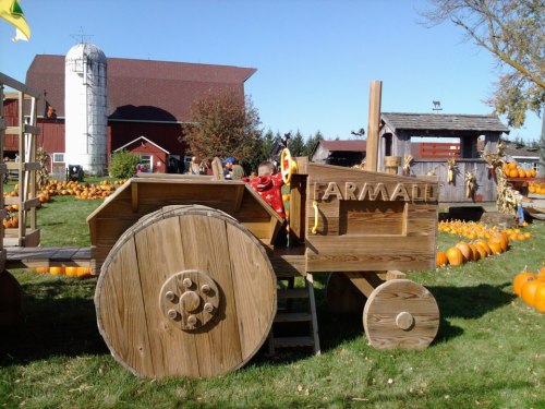 A wooden tractor play structure in a pumpkin patch, with children playing and a barn in the background.