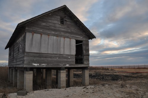 Abandoned wooden house on stilts, set against a cloudy sky and barren landscape.