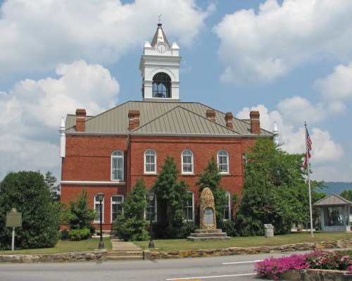 Historic brick courthouse with a clock tower, surrounded by greenery and flowers, under a partly cloudy sky.