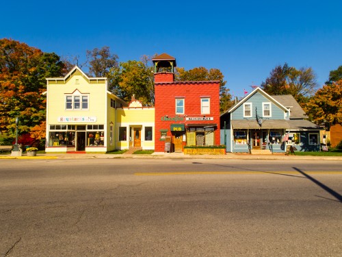 Colorful storefronts line a street, featuring a yellow shop, a red pub, and a blue house, with autumn trees in the background.