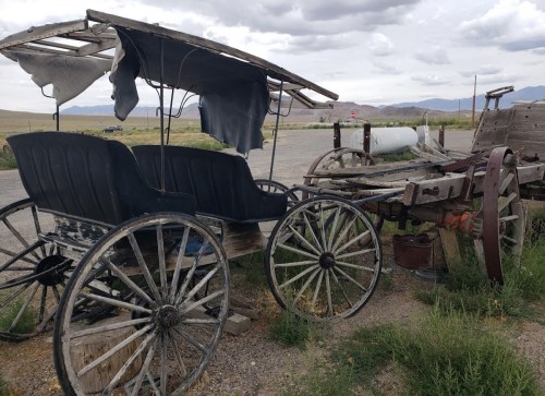 An old, weathered horse-drawn carriage sits beside a rustic wagon in a grassy area under a cloudy sky.
