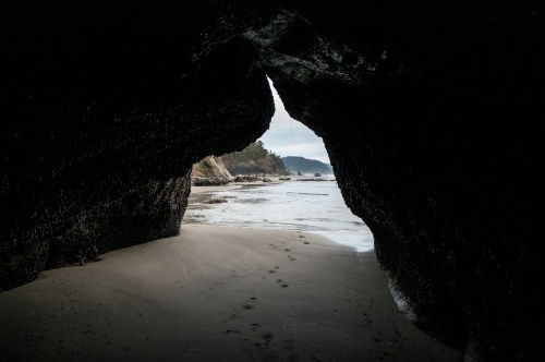 View from a cave opening onto a sandy beach and calm ocean, with footprints leading towards the water.