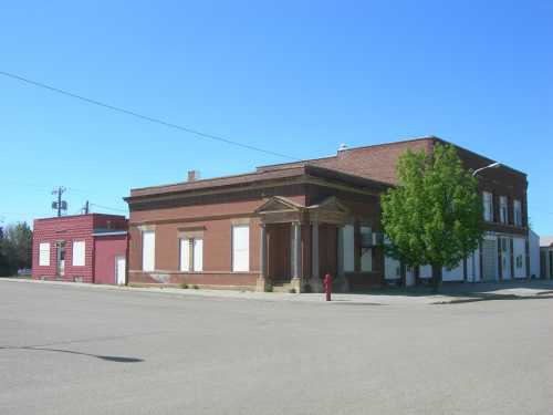 A brick building with a columned entrance, situated on a street corner, with a smaller red building nearby. Clear blue sky.