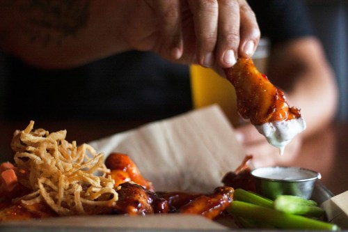 A hand dips a chicken wing into ranch dressing, surrounded by celery sticks and crispy onions.