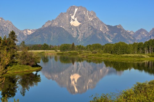 A serene mountain landscape with a clear blue sky, reflecting in a calm lake surrounded by lush greenery.
