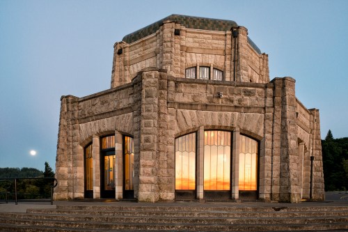 A stone building with large windows reflecting a sunset, set against a twilight sky and distant trees.