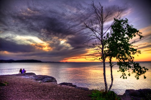 A serene sunset over a lake, with a couple sitting on the shore beside a lone tree against a colorful sky.