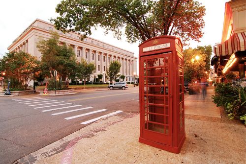 A red telephone booth stands on a sidewalk near a street, with a historic building and trees in the background at sunset.