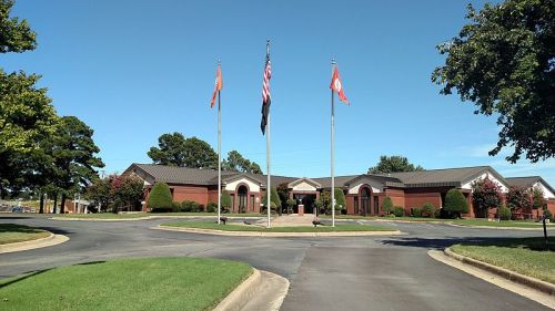 A brick building with flags in front, surrounded by trees and a circular driveway under a clear blue sky.